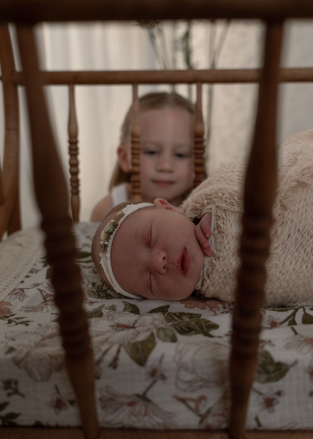 Big sister smiling at sleeping newborn baby in a vintage wooden crib with floral bedding.