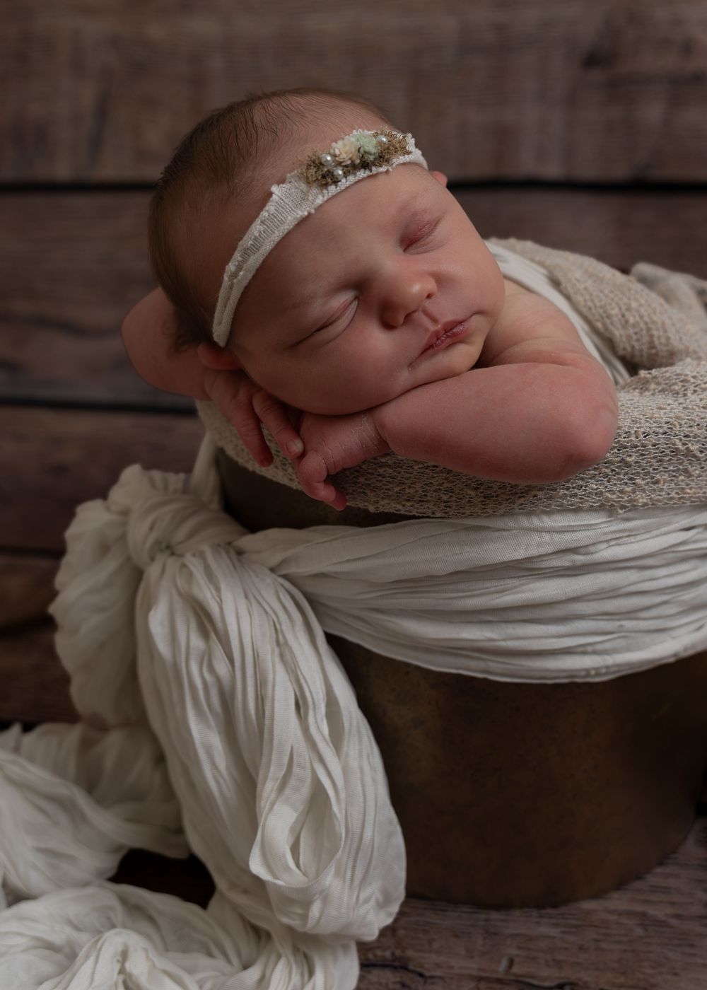 Sleeping newborn baby posed in a bucket with cream wrap and floral headband.