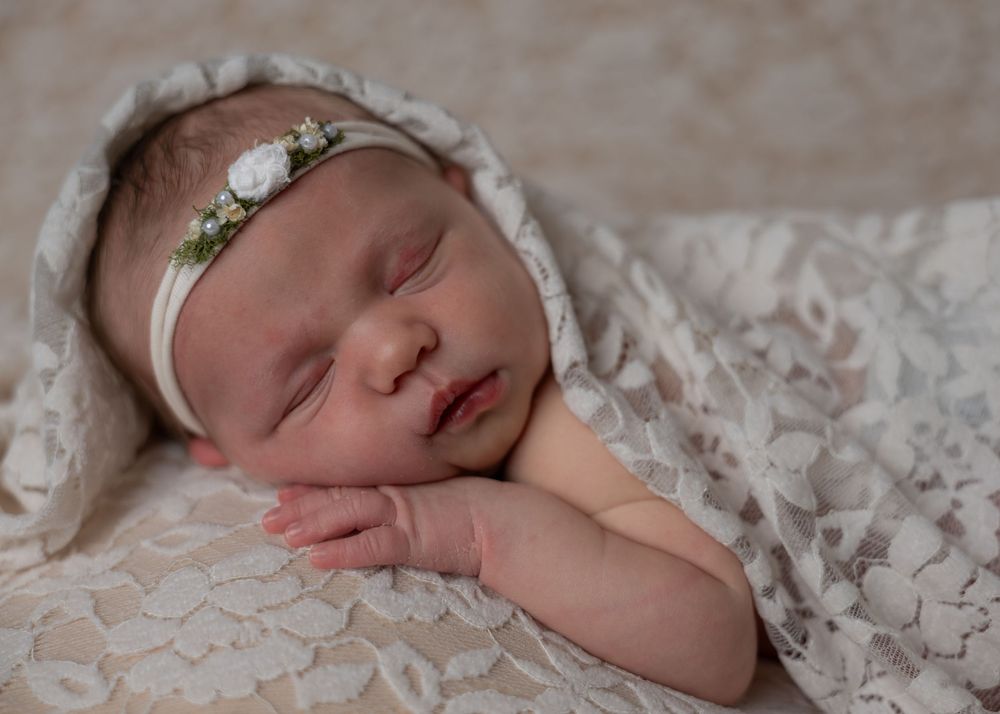Peaceful newborn baby girl wrapped in white lace with a small floral headband.