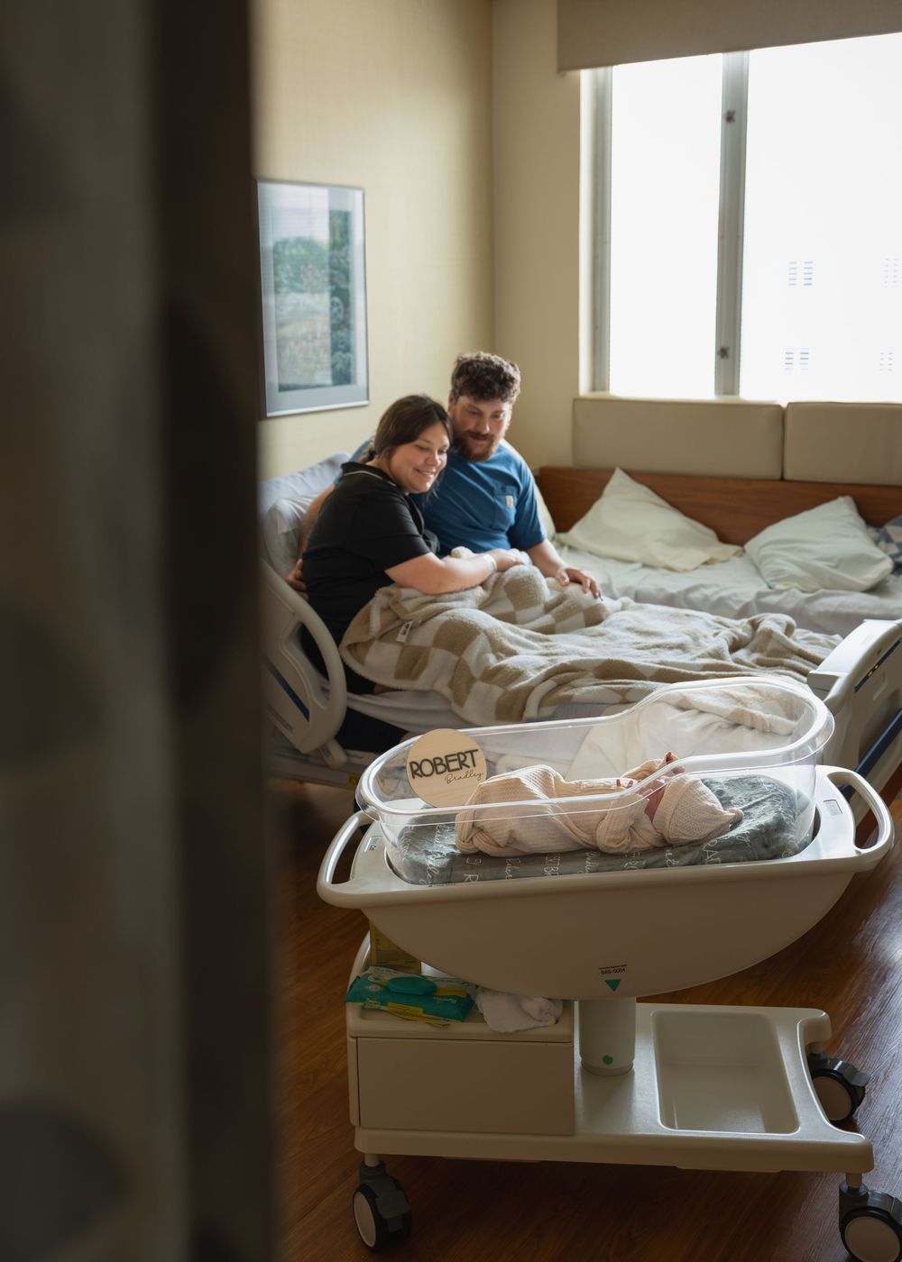New parents admire their newborn baby named Robert in a hospital room bassinet after delivery.