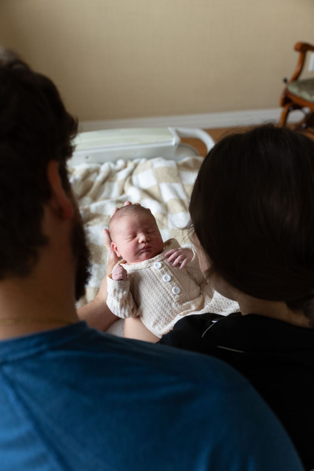 Parents gazing lovingly at their newborn baby dressed in a white knit outfit, lying peacefully in their arms.