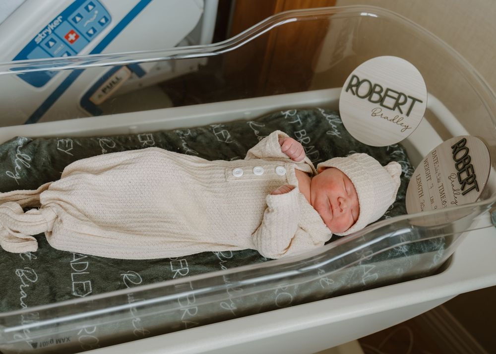 Newborn baby in hospital bassinet wearing a white gown and hat after birth.