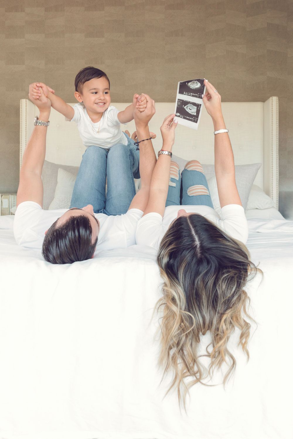 Family lying on bed holding ultrasound photo in pregnancy announcement pose.