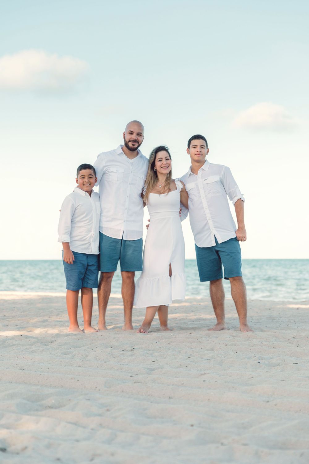 Family group poses together on beach sand at sunset wearing coordinated blue and white outfits.