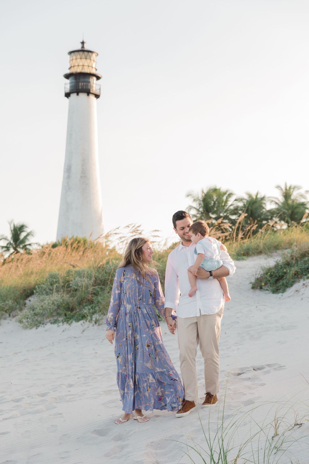 Family walks along beach path near white lighthouse surrounded by palm trees at sunset.
