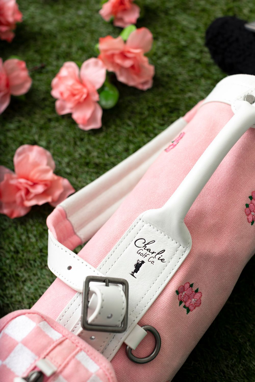 Pink golf bag with white strap, Charlie and Co. branding, floral embroidery, on green grass with pink flowers.
