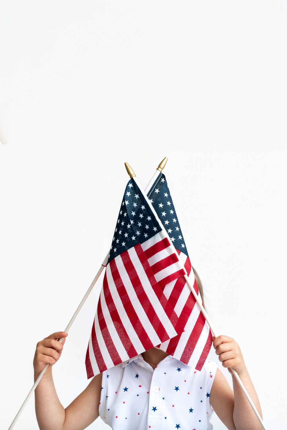 Child in star-print shirt holding two crossed American flags in front of face on white background.