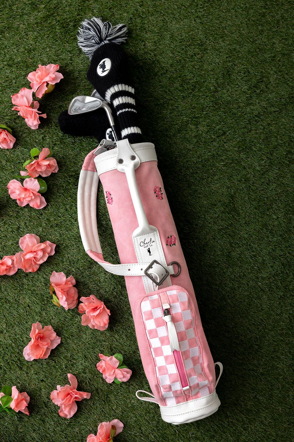 Pink and white checkered golf bag with clubs surrounded by coral flowers on artificial grass background.