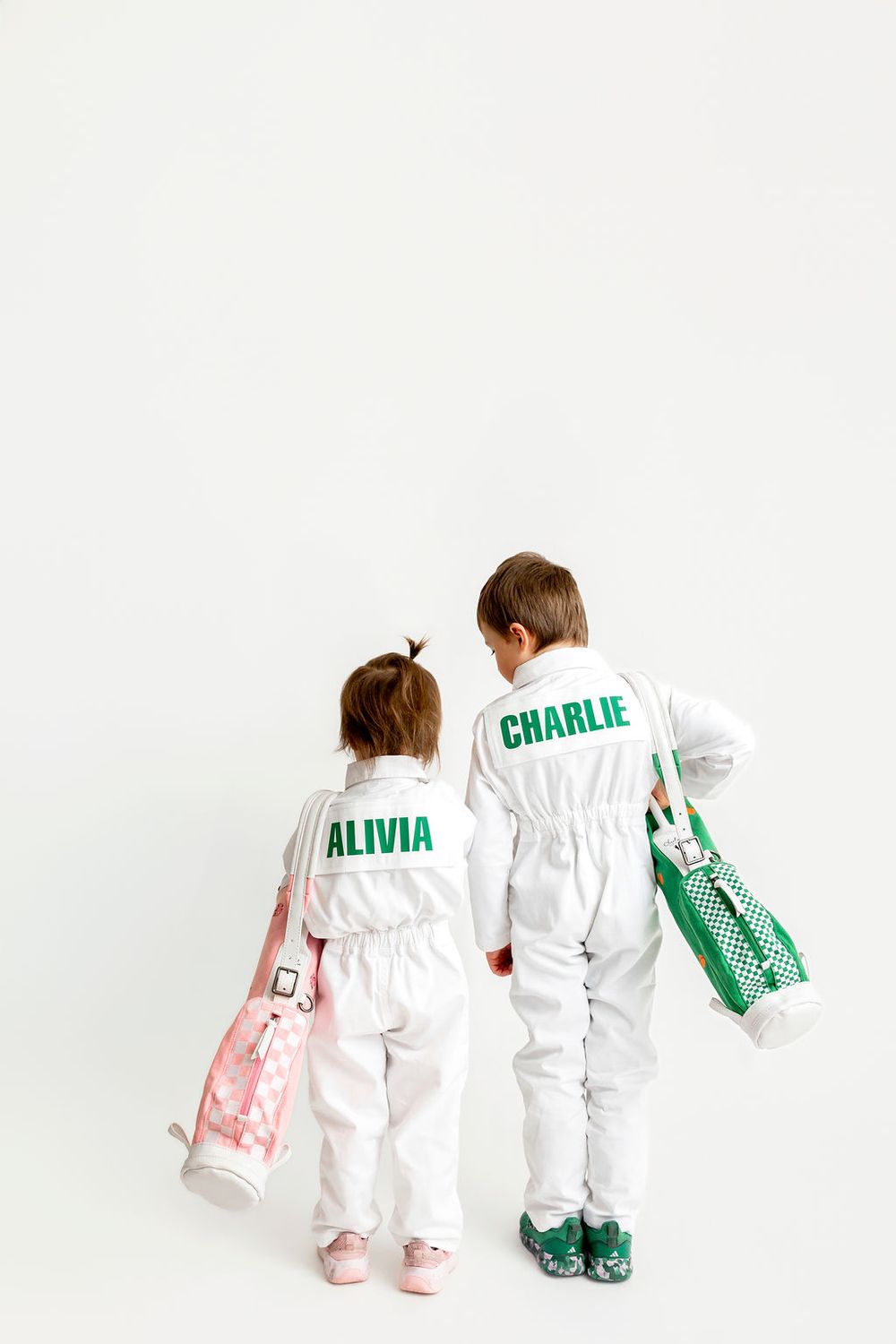 Two children in matching white coveralls with personalized names walk together holding bags and wearing colorful shoes.