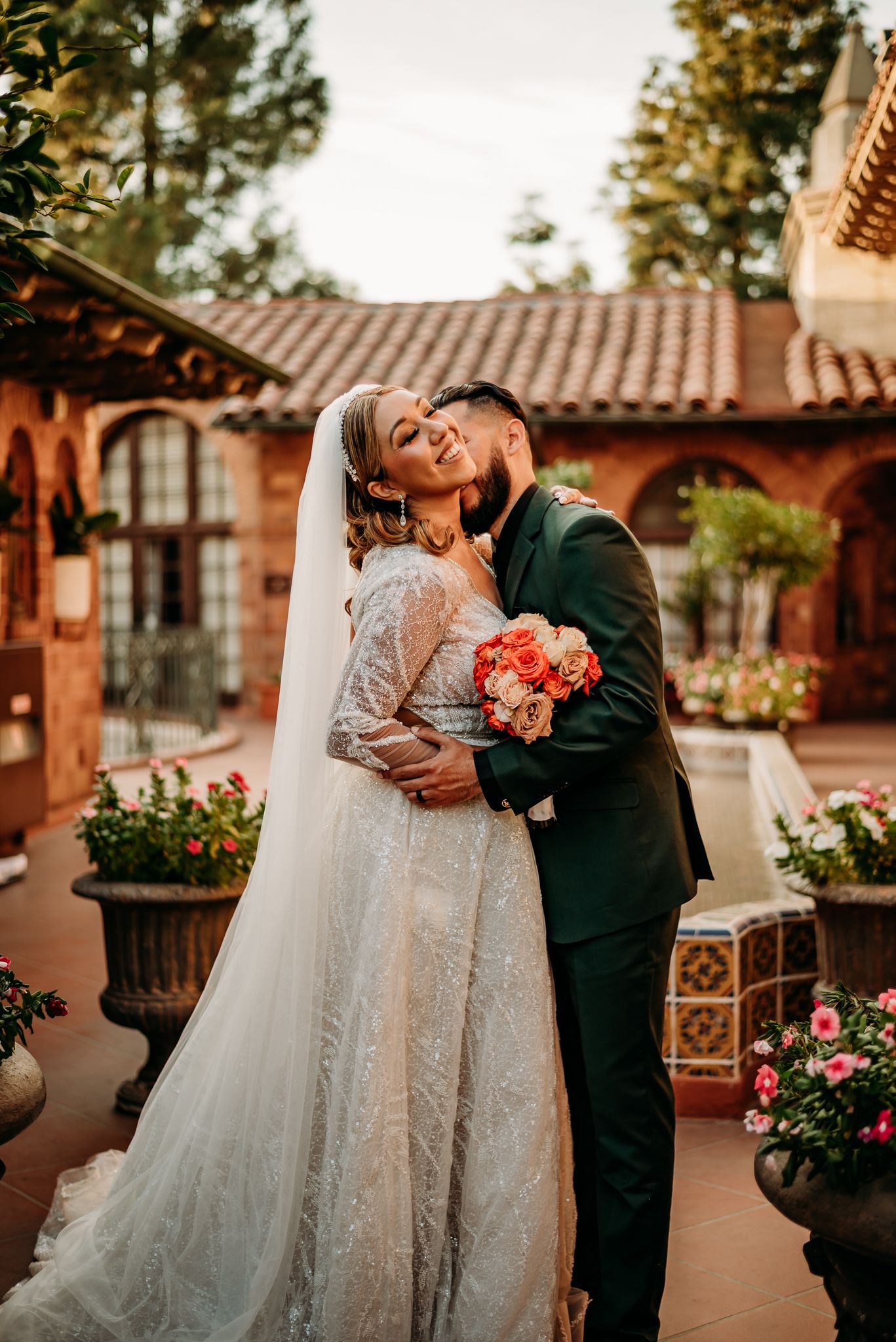 A bride and groom share a romantic moment among lush trees, she in a lace gown with veil, he in a blue suit.