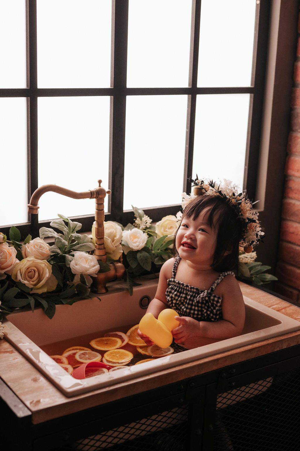 Smiling toddler girl wearing a floral crown sits in a decorative sink with orange slices and roses.
