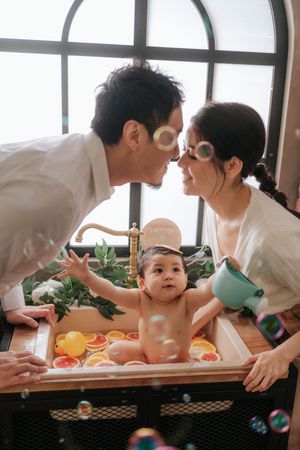 A couple shares a tender moment leaning in for a kiss over their baby sitting in a decorative fruit bath with bubbles.