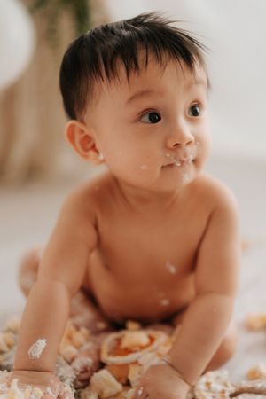 Adorable baby with cake smeared on face sitting on floor during first birthday cake smash photoshoot.