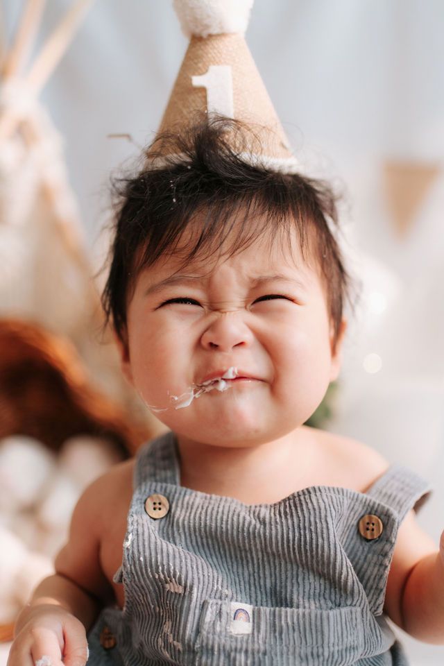 Cute baby celebrating first birthday wearing a number one party hat with cake on face, smiling joyfully.