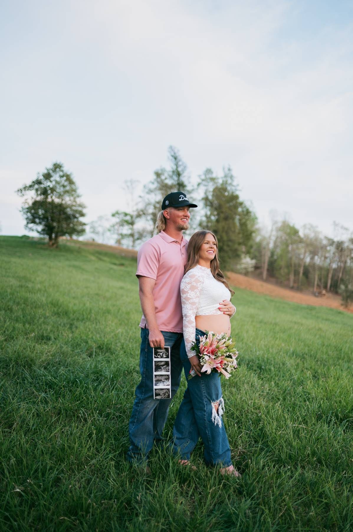 Couple standing in green field holding ultrasound photos and flowers, announcing pregnancy at golden hour in North Carolina