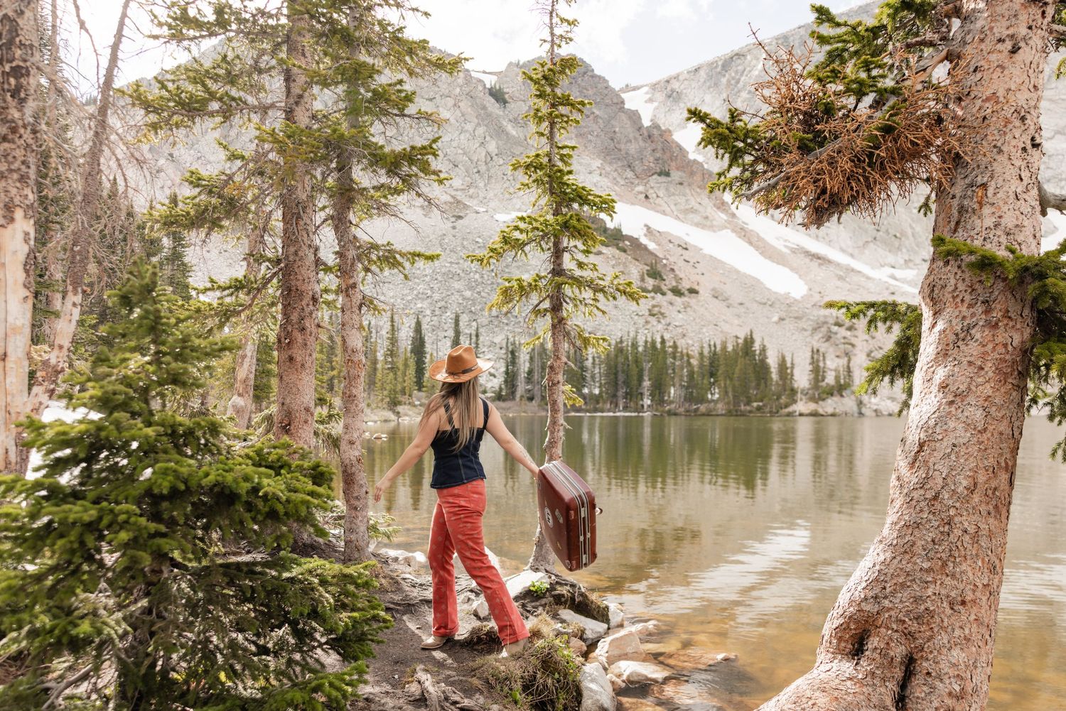Woman in red pants and hat stands by a serene mountain lake surrounded by pine trees and rocky peaks.