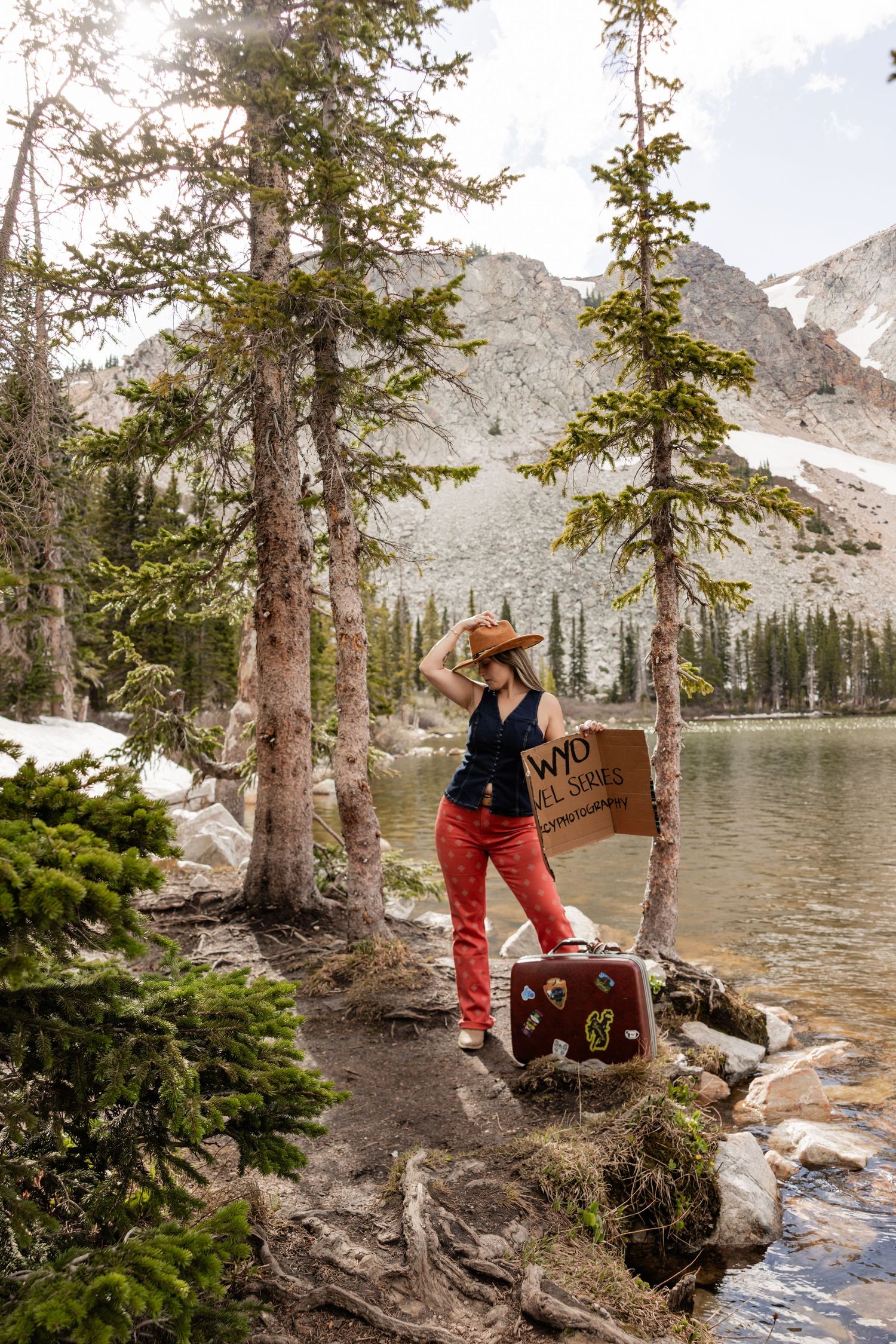 Woman in red pants and cowboy hat stands by mountain lake with vintage suitcase and Wyoming sign among pine trees.