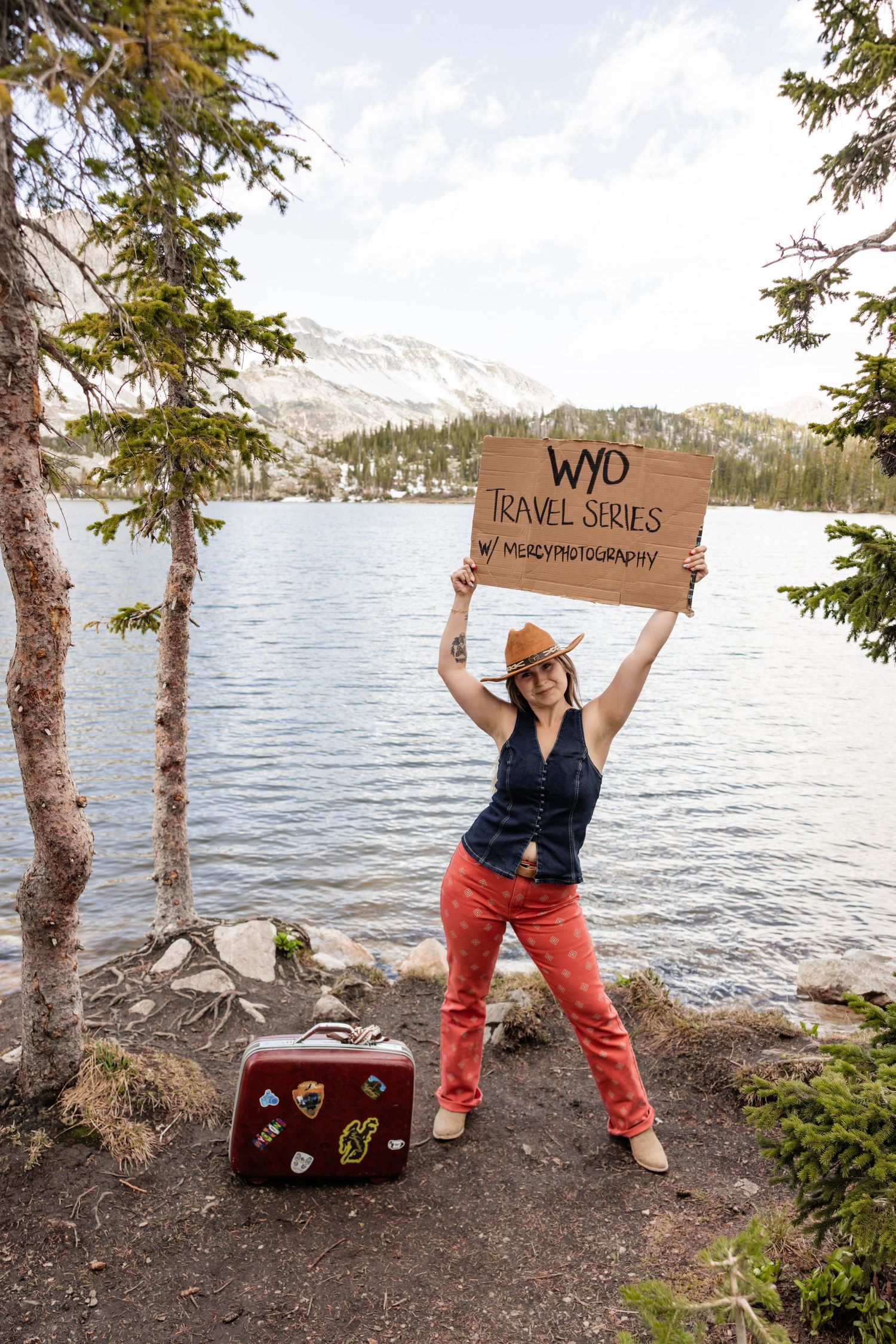 Woman holding WYD Travel Series sign by mountain lake with vintage sticker suitcase, wearing red pants and cowboy hat.