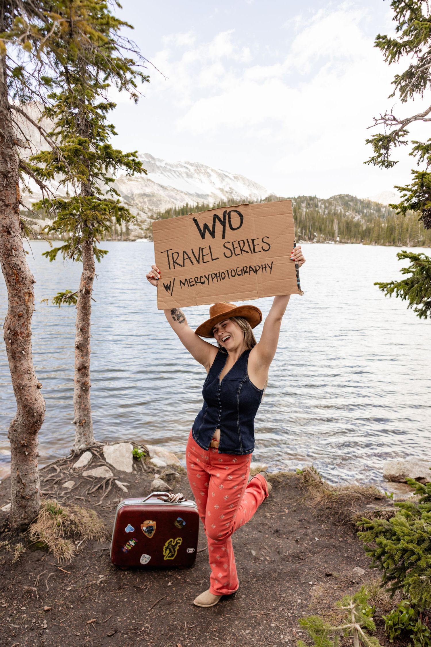 Woman holding WYD Travel Series sign by a mountain lake, wearing a cowboy hat and red pants with a vintage suitcase.