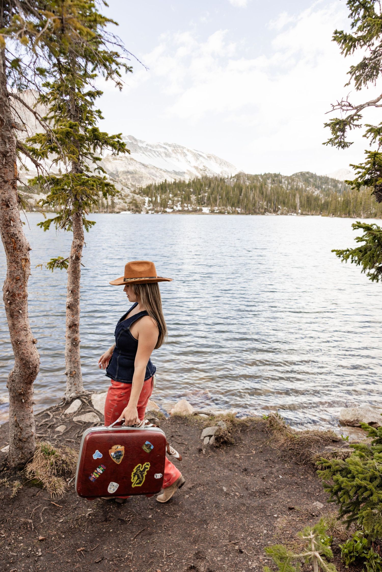 Woman in brown hat carrying vintage sticker-covered suitcase along scenic mountain lake shore surrounded by pine trees.