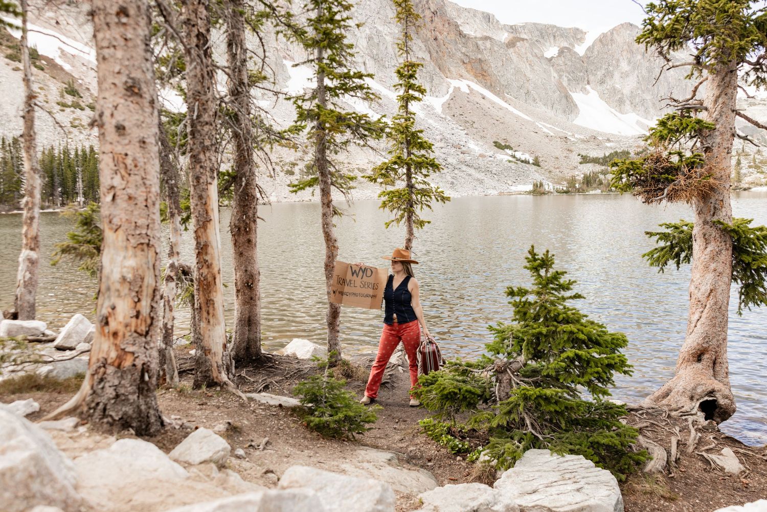 Hiker in red pants and hat walks along a scenic alpine lake trail surrounded by pine trees and rocky mountains.