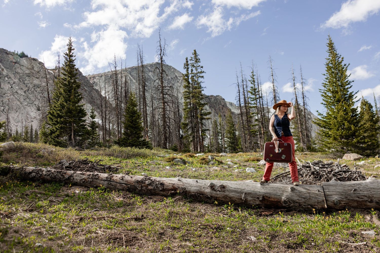 Woman hiking on fallen logs in a mountain forest with rocky peaks and evergreen trees under a blue sky.