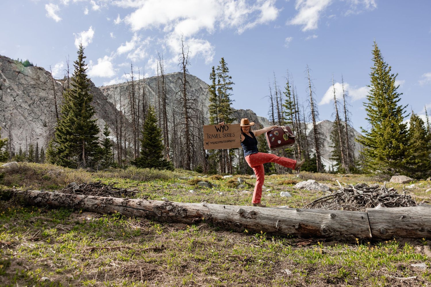 Woman balancing on a fallen log in a mountain forest, holding a sign while wearing red pants on a sunny day.