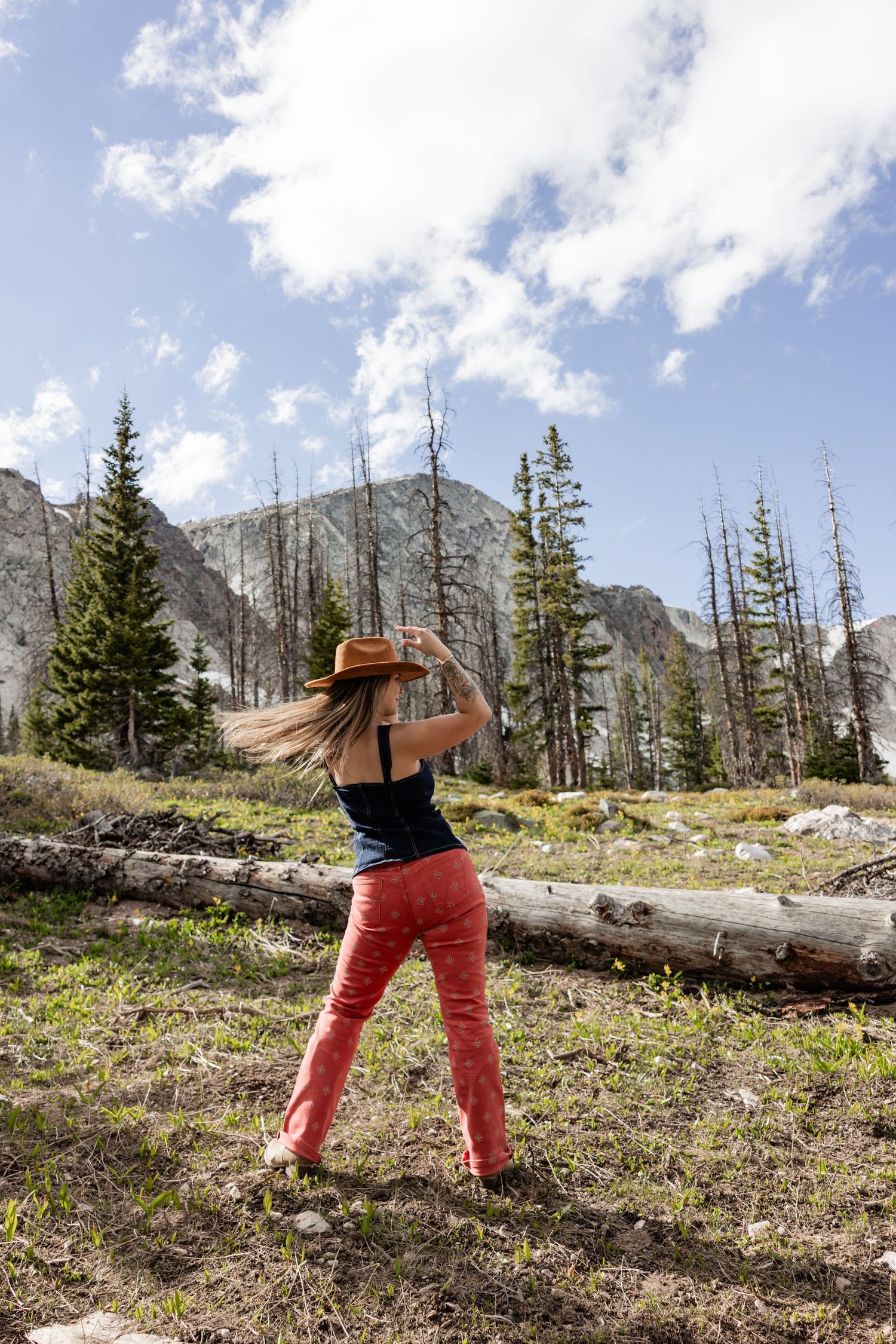 Woman in red pants and cowboy hat poses in mountain meadow surrounded by pine trees under a partly cloudy sky.