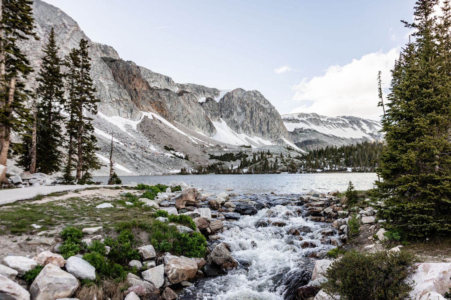 A rushing mountain stream flows past rocky shores toward a serene alpine lake beneath snow-dusted granite peaks.