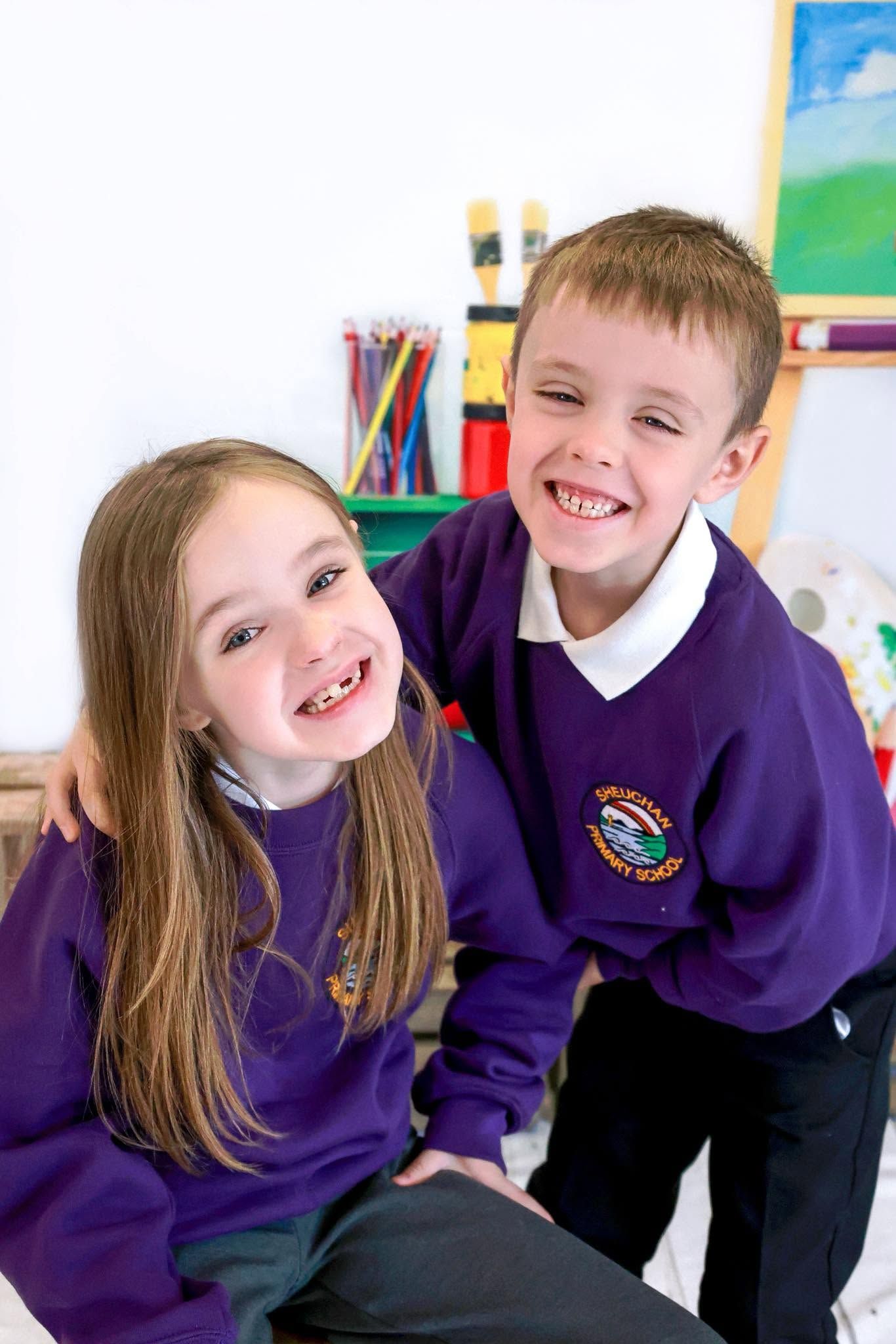 Two smiling children in purple school uniforms posing together in a colorful classroom setting.