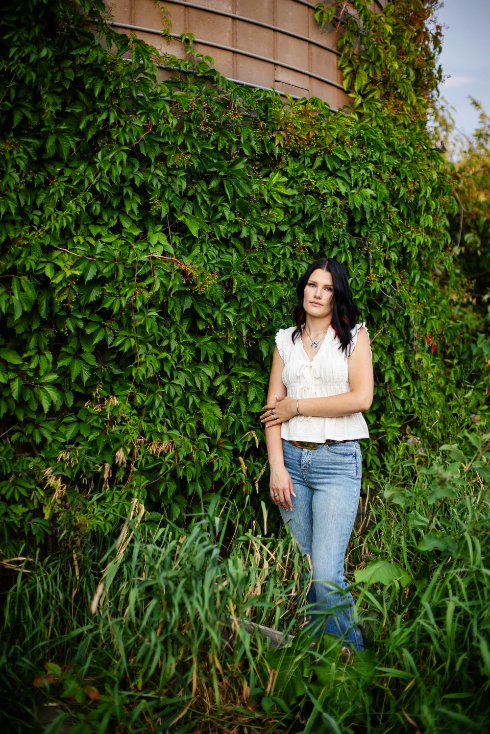 Woman in white top and jeans standing against ivy-covered brick wall surrounded by lush green foliage outdoors.