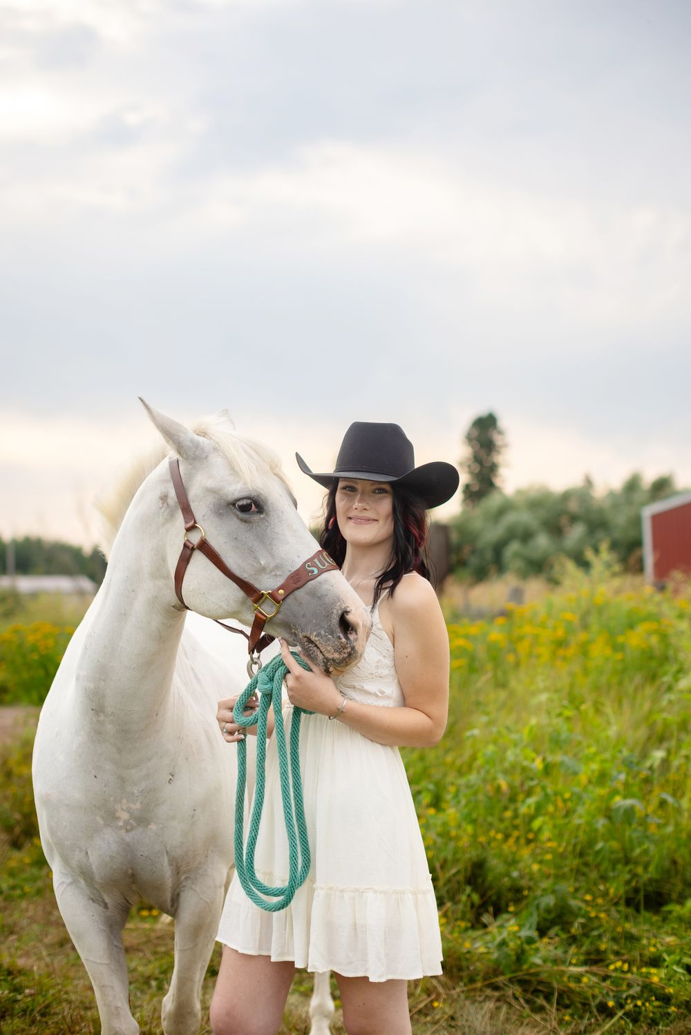 Young woman in black cowboy hat and white dress stands beside a white horse in a green field.