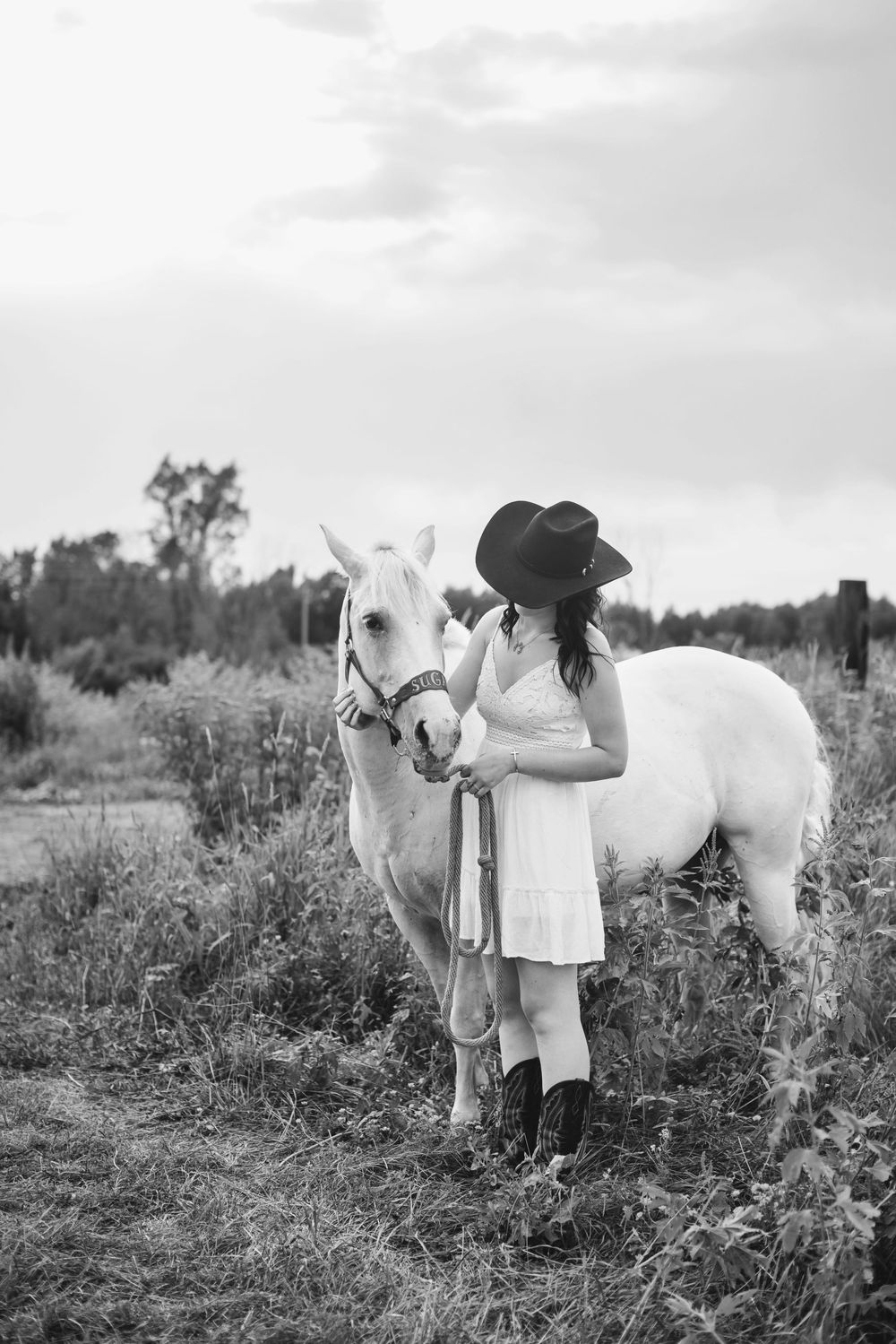 Black and white photo of a woman in a cowboy hat and white dress standing beside a white horse in a field.