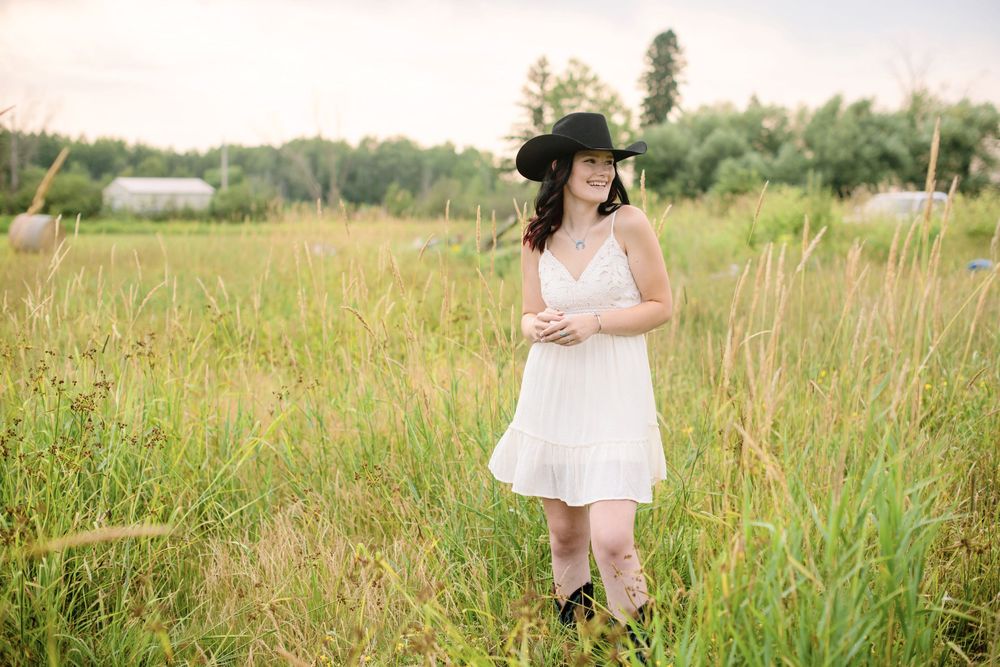 Woman in white dress and black hat standing in a lush green field during golden hour.