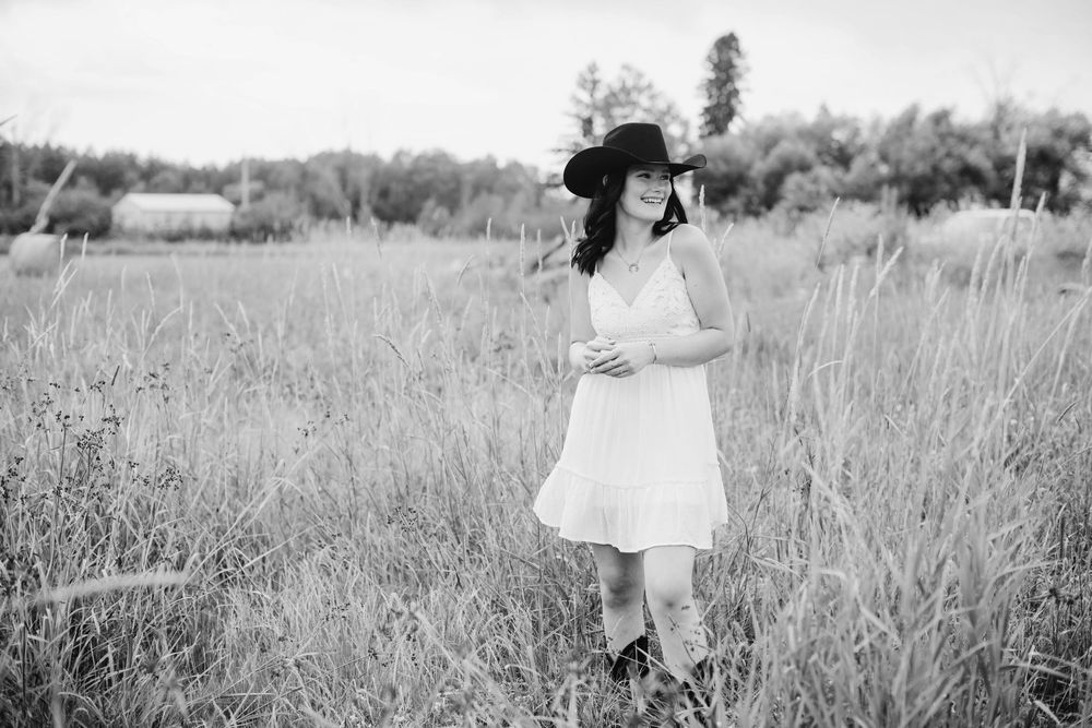 Woman in white dress and black hat standing in tall grass field, black and white outdoor portrait photography.