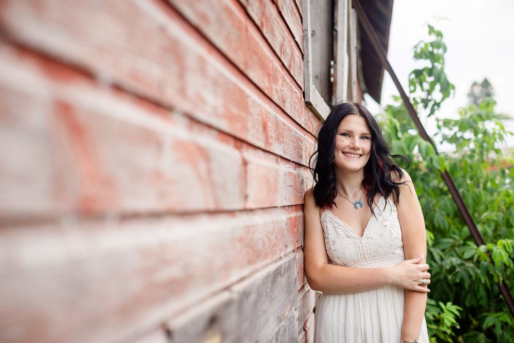 Young woman in white dress leaning against rustic red brick wall outdoors, smiling with dark hair and necklace.