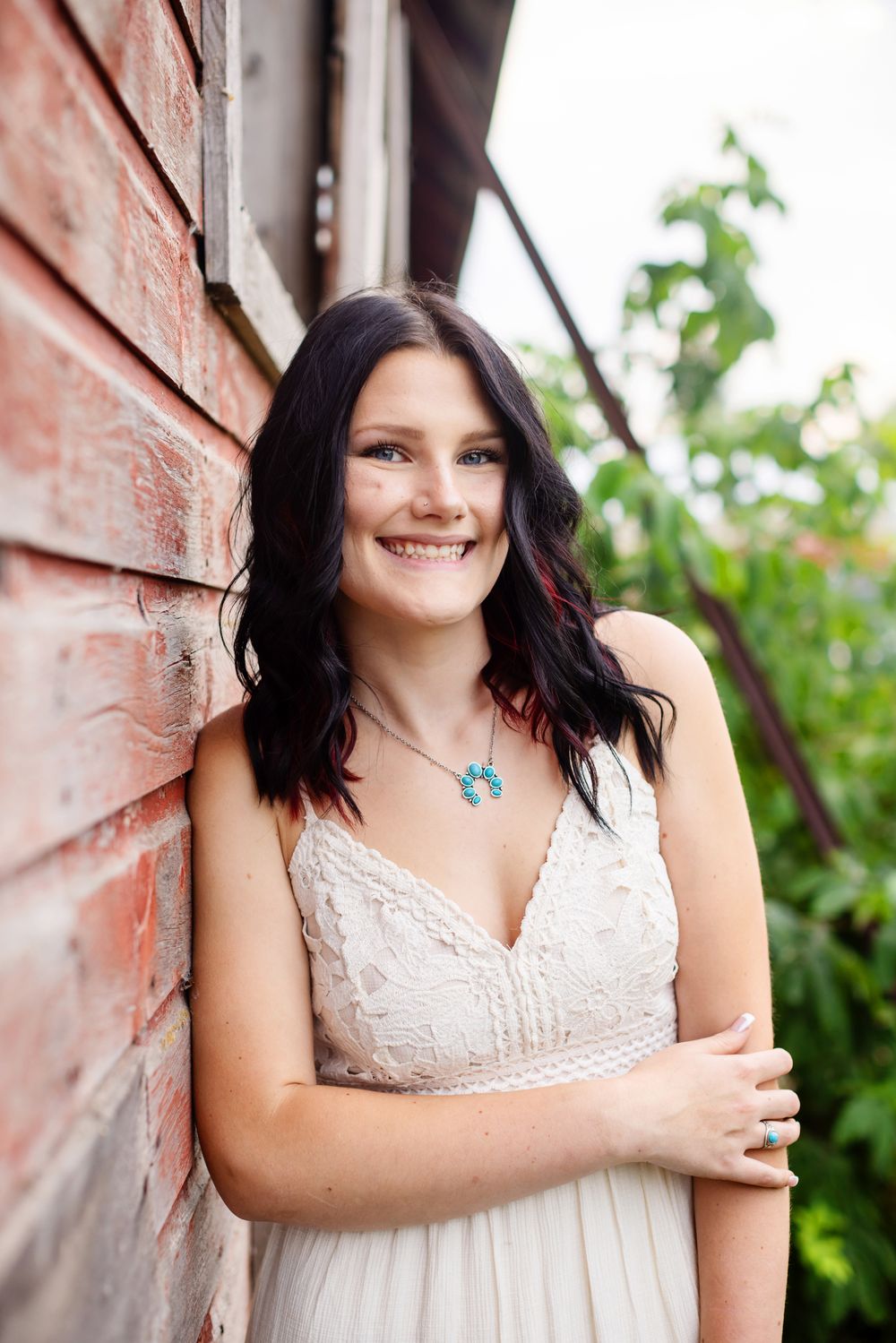 Smiling young woman with dark hair in white lace dress leaning against brick wall outdoors wearing turquoise necklace.