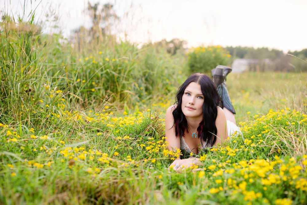 Young woman with dark hair lying in a field of yellow wildflowers, gazing thoughtfully into the distance.