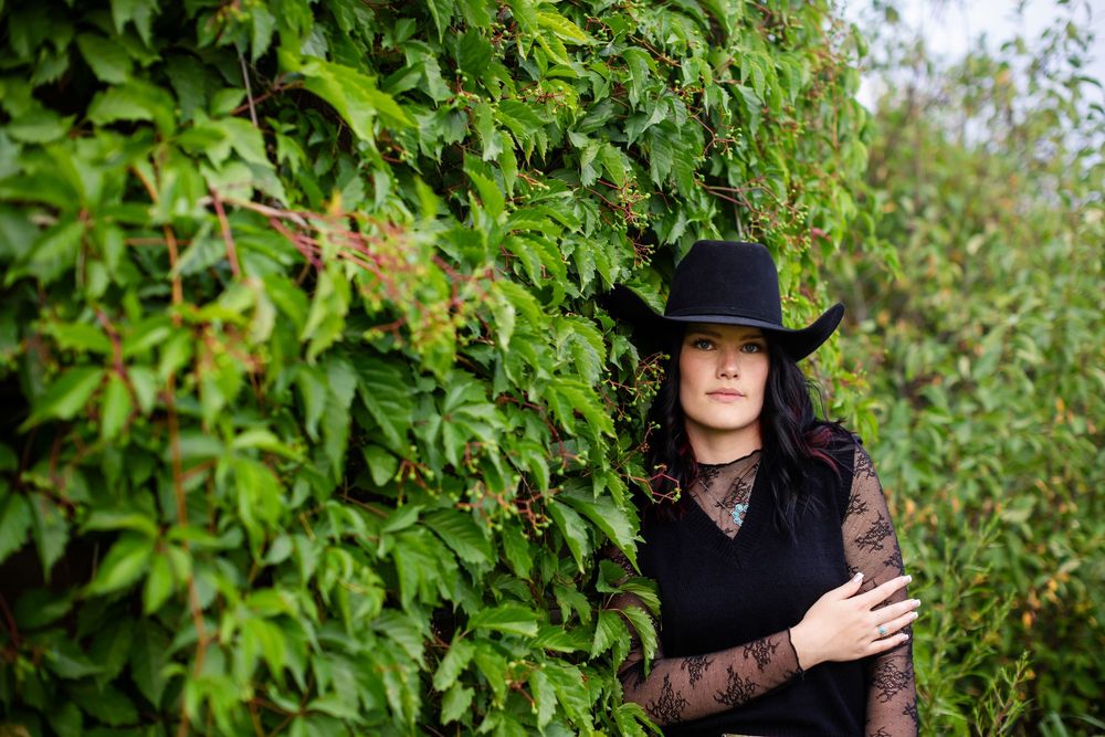 Woman in black cowboy hat and lace top posing against lush green ivy wall outdoors.