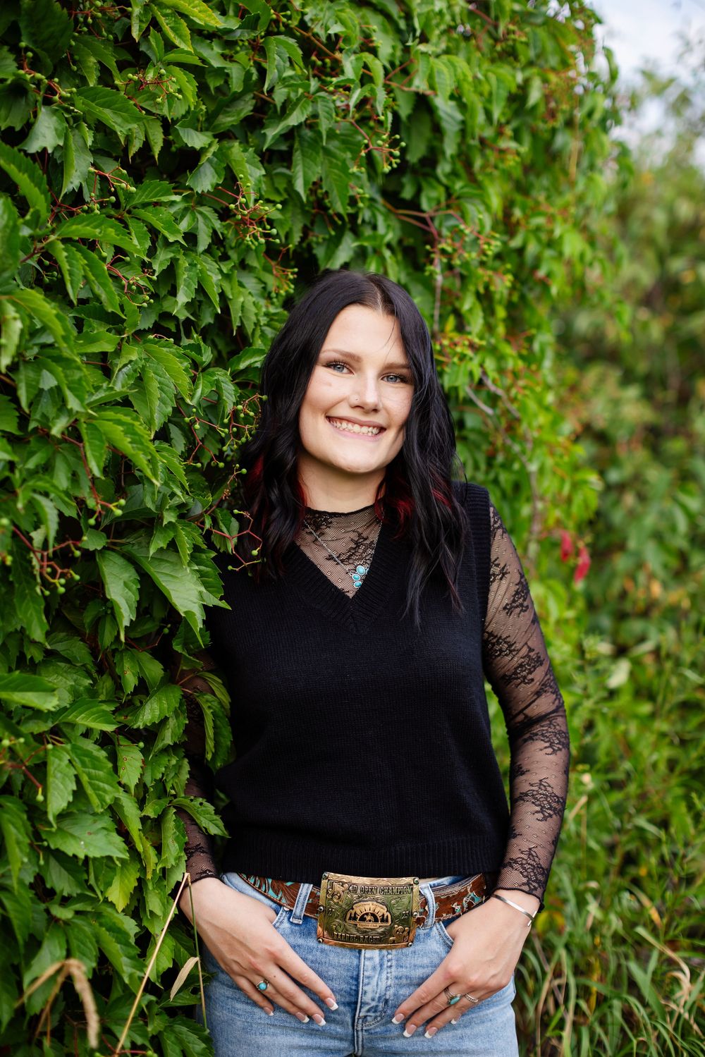 Young woman with dark hair smiles outdoors, wearing black vest, lace sleeves, jeans, and decorative belt buckle.