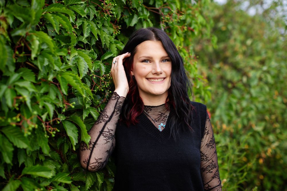 Smiling woman with dark hair in black lace top posing outdoors against lush green ivy wall.