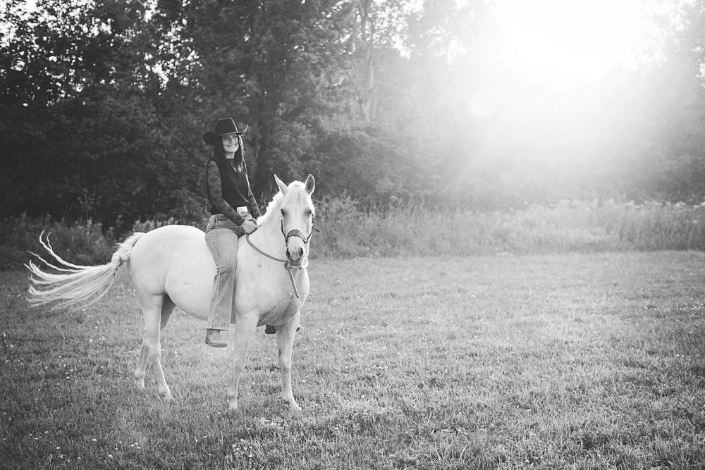Black and white photo of a person riding a white horse in a grassy field with trees in the background.