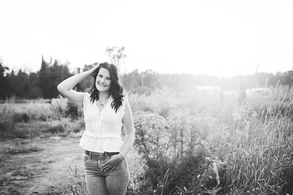 Black and white photo of a smiling young woman in a white top and jeans posing in a sunlit field.