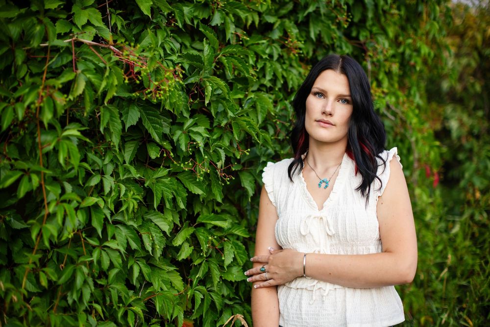 Woman in white top with turquoise jewelry poses before lush green ivy wall outdoors.