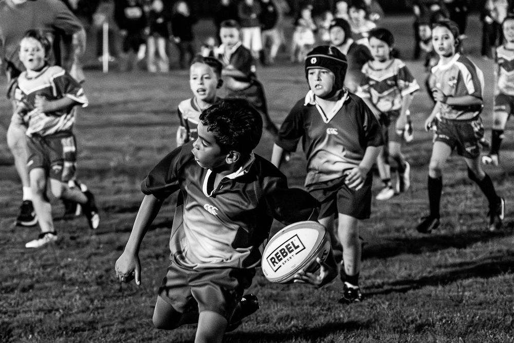 Black and white photo of young children playing rugby, with one player carrying the ball during an intense match.