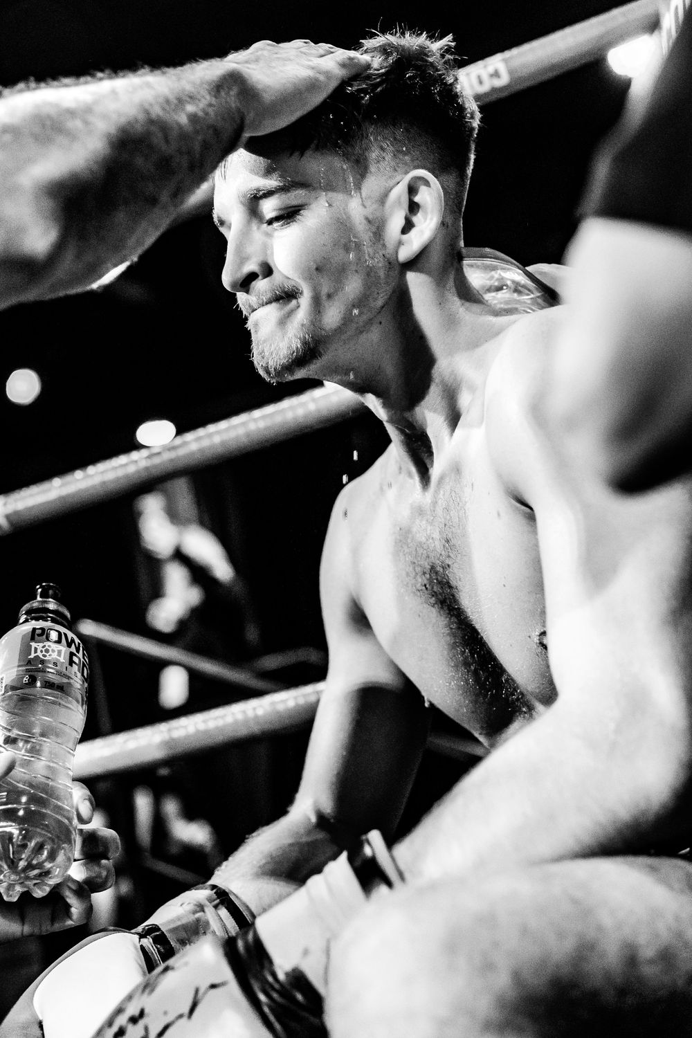 Black and white photo of a sweaty shirtless boxer resting in the corner of a boxing ring with a water bottle.