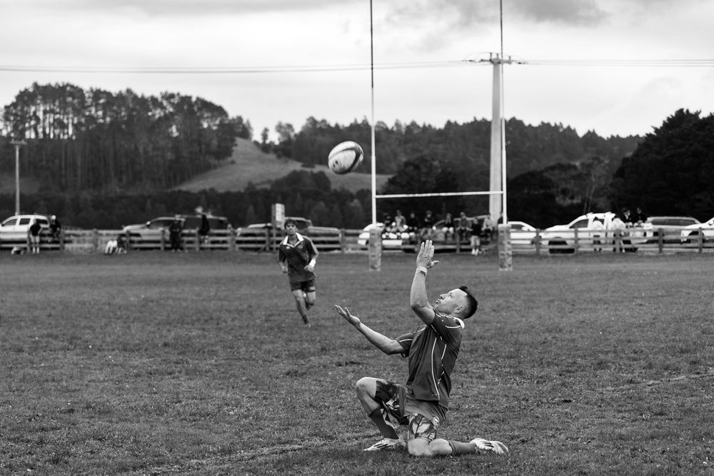 Black and white photo of rugby players on a field, one leaping to catch a ball mid-air during a match.