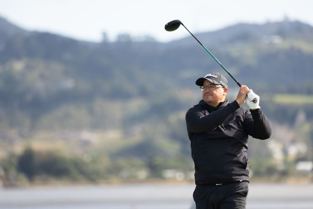 Golfer in black outfit swinging a driver on a scenic coastal golf course with mountains in the background.