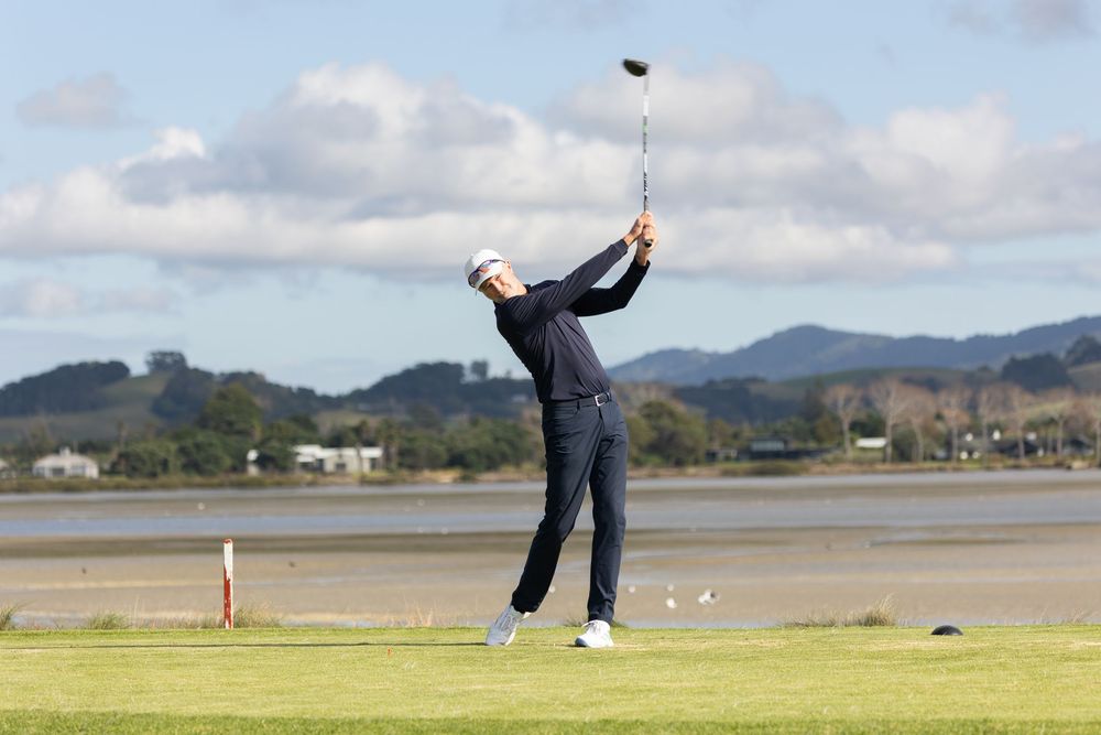 Golfer completing a swing on a scenic coastal golf course with hills and water in the background.