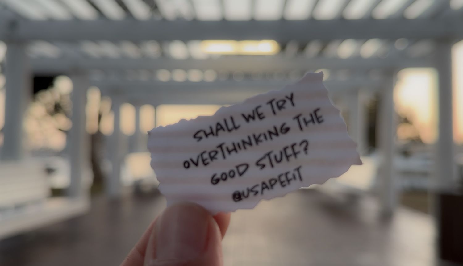 Hand holding a torn paper note reading 'Shall we try overthinking the good stuff?' against blurred pergola backdrop.